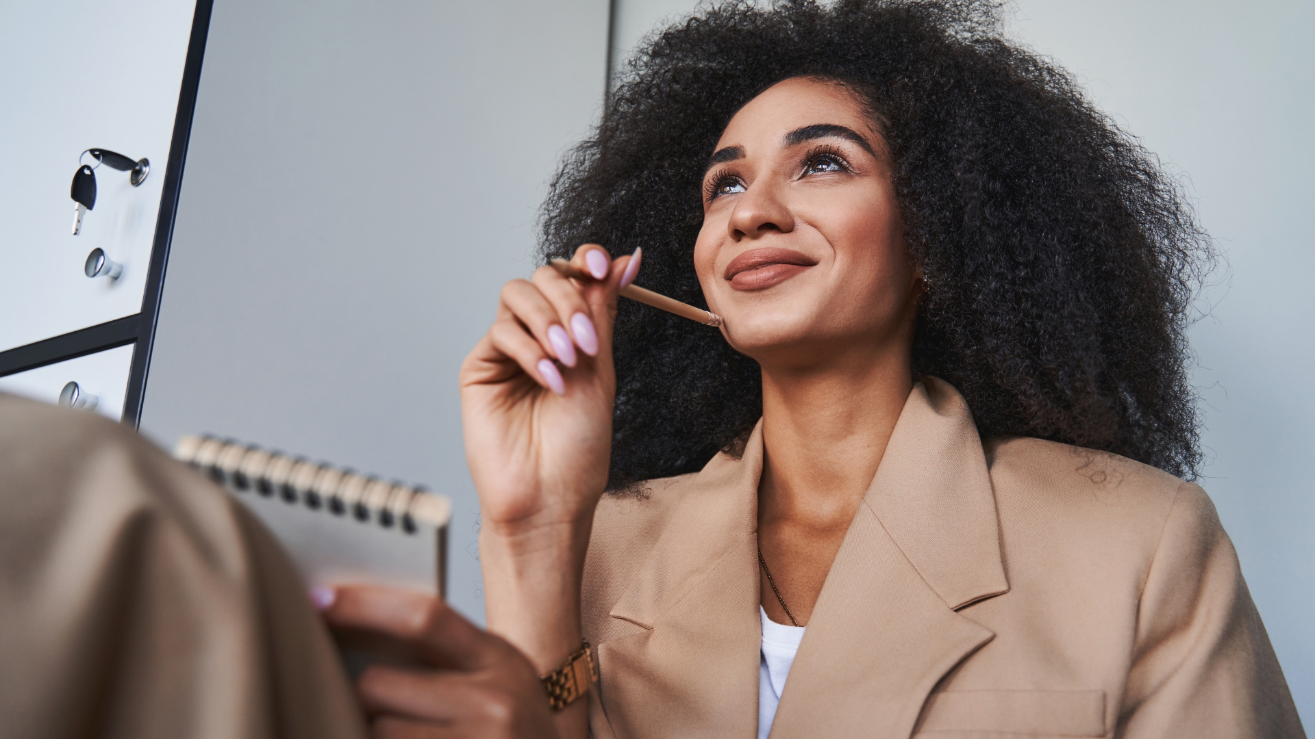 Mulher sorrindo e pensativa segurando um lápis e um bloco de notas, refletindo sobre planejamento de carreira e objetivos futuros